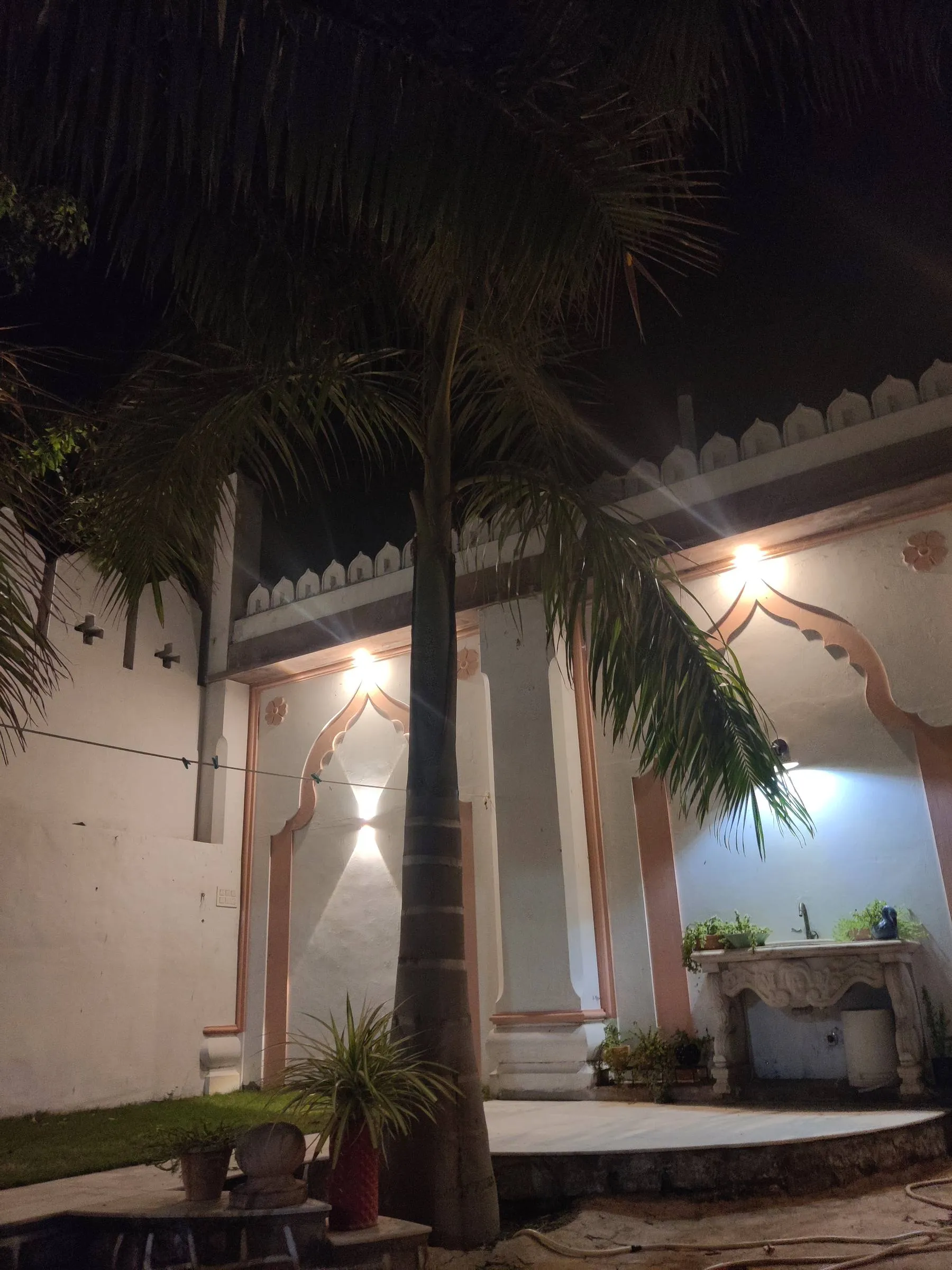The courtyard at Rawla Bankli after dark, wall lamps washing the white walls behind young palms