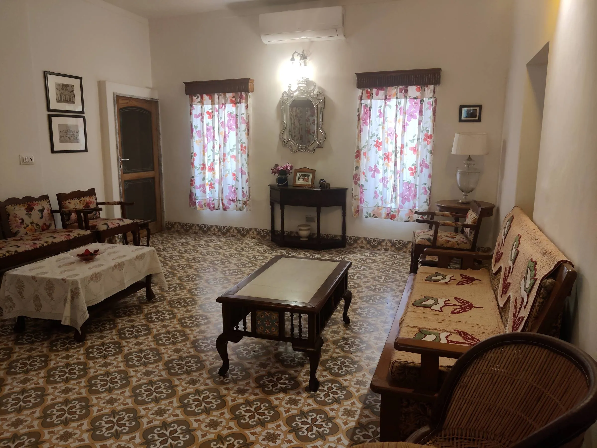 Afternoon sun across the sitting room's geometric tiled floor, floral curtains drawn back at two tall windows